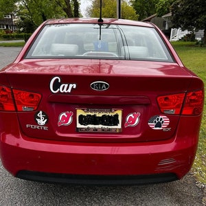 May include: A red Kia Forte car with a "Car" sticker on the back. The car has a New Jersey license plate and several other stickers, including a hockey team logo, an anchor, and a paw print.