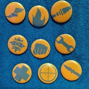 May include: Nine yellow round buttons with blue graphics. The graphics include a winged shoe, a flame, a rocket, a spiked ball, a fist, a wrench, a cross, a target, and a knife. The buttons are arranged on a blue fabric surface.