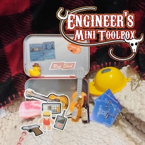 May include: A silver metal tin labeled Red Shed containing miniature items including a yellow rubber duck, a guitar, two small bottles, a yellow hard hat keychain, and various stickers featuring a bear, a handgun, and blueprints.