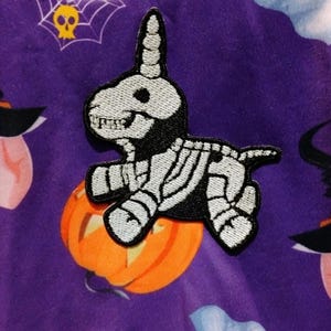 Può includere: Toppa ricamata con un unicorno scheletro su una zucca arancione. La toppa presenta dettagli in bianco e nero, su uno sfondo viola decorato con motivi di Halloween, tra cui una ragnatela e un cappello da strega.