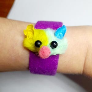 May include: A purple wristband with a colorful, fluffy mouse face. The mouse has yellow, white, and blue fur, black eyes, and a pink nose. The wristband is made of a soft, fuzzy material.