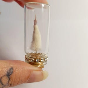 May include: A small, clear glass vial containing a miniature, fluffy, cream-colored tree. The base of the vial is adorned with a decorative gold and rhinestone band. The tree appears to be handmade and delicate.