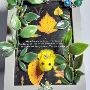 May include: A framed artwork with a yellow mouse figure and decorative green leaves. The frame is gray, and the artwork features a black background with yellow leaves and text that reads "Miracles are in the air you breath..."