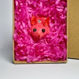 May include: A pink rodent figurine with a jeweled crown, nestled in a box filled with bright pink shredded paper. The box is a natural cardboard color, and the rodent has large black eyes and orange ears.