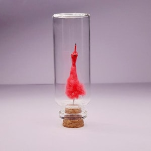 May include: A clear glass bottle with a cork base, containing a small, vibrant red, feathery dress. The dress is suspended inside the bottle, creating a unique decorative piece. The background is a soft, gradient purple.