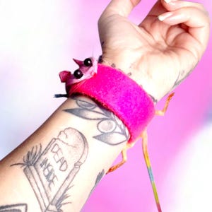 May include: A quirky pink mouse bracelet with a rainbow tail. The bracelet is a bright fuchsia colour and wraps around the wrist. The mouse has large black eyes, a small nose and ears.