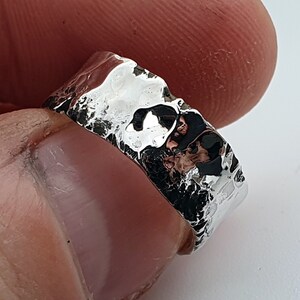 Puede incluir: Un anillo de color plateado con una banda ancha y una superficie martillada irregular altamente reflectante. El acabado metálico es pulido y brillante, creando contrastes claros y oscuros definidos en todo el exterior irregular.