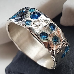 May include: A silver ring with a textured surface and inlaid with blue gemstones. The ring has a wide band and a unique, organic design. The gemstones are round and evenly spaced around the ring.