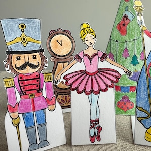 Peut inclure: Découpages de personnages dessinés à la main du ballet Casse-Noisette. Les personnages comprennent un soldat casse-noisette, une horloge, une ballerine en tutu rose, un sapin de Noël et le Roi des Souris. Les personnages sont colorés aux marqueurs.
