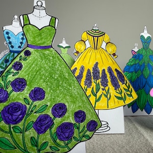 Puede incluir: Una colección de coloridos vestidos de muñecas de papel en exhibición. Los vestidos están dibujados a mano y coloreados con rotuladores, con varios diseños, incluyendo patrones florales y detalles en forma de hojas. Cada vestido está montado en un maniquí de papel.