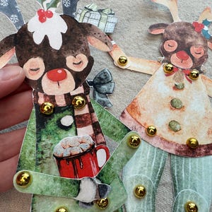 Op de afbeelding: Papieren kerstrendierfiguren met beweeglijke ledematen, met aquarel illustraties. Een rendier draagt een groene jas en sjaal, met een mok warme chocolademelk. Het andere rendier heeft een peperkoekachtig ontwerp. Gouden brads verbinden de ledematen.