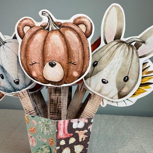 Puede incluir: Una colección de recortes de papel con temática otoñal, que incluyen un zorro, un oso y un conejo, cada uno diseñado como una calabaza. Los recortes están dispuestos en un recipiente decorativo con un patrón otoñal.