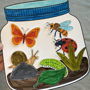 Peut inclure: Une découpe en papier en forme de bocal avec un couvercle bleu, contenant des illustrations d'un papillon, d'une abeille, d'une coccinelle, d'un escargot, d'une chenille verte, d'une pierre grise, d'une feuille verte et d'herbe.