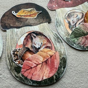 Könnte beinhalten: Aquarellillustrationen von pilzförmigen Ausschnitten mit Waldszenen. Jeder Ausschnitt zeigt ein schlafendes Eichhörnchen, Blätter und Eicheln. Die Farbpalette umfasst Brauntöne, Rot und Grün.