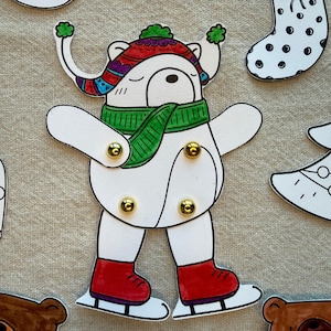 Puede incluir: Un recorte de papel blanco de un oso con un sombrero colorido, una bufanda verde y patines de hielo rojos. El oso tiene sujetadores dorados en las articulaciones. Tambi&eacute;n se ven otros recortes con temas navide&ntilde;os.