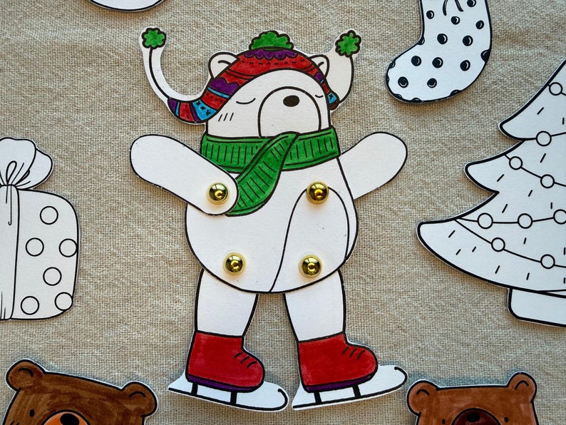 Puede incluir: Un recorte de papel blanco de un oso con un sombrero colorido, una bufanda verde y patines de hielo rojos. El oso tiene sujetadores dorados en las articulaciones. Tambi&eacute;n se ven otros recortes con temas navide&ntilde;os.