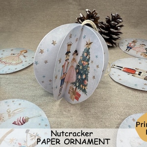 Può includere: Decorazione di carta di Natale con motivo di Schiaccianoci stampabile. L'ornamento presenta uno Schiaccianoci e una ballerina in piedi davanti a un albero di Natale. L'ornamento è realizzato in carta ed è progettato per essere piegato e assemblato.