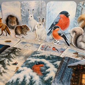 May include: A selection of winter-themed paper cutouts, including a fox, squirrel, and birds, arranged on illustrated cards. The cards depict winter scenes and text. The colour scheme features white, brown, and red.