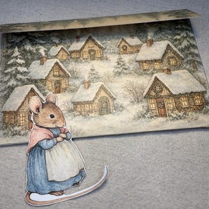 May include: A whimsical illustration of a mouse in a blue dress and pink shawl, standing on a curved base. Behind the mouse is a winter scene with snow-covered houses and trees, creating a cozy, festive atmosphere.
