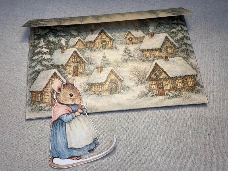 May include: A whimsical illustration of a mouse in a blue dress and pink shawl, standing on a curved base. Behind the mouse is a winter scene with snow-covered houses and trees, creating a cozy, festive atmosphere.