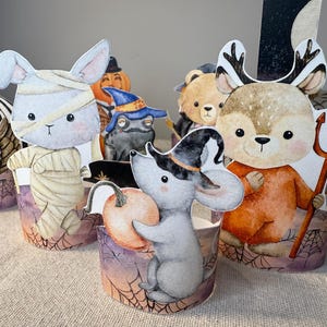 May include: A collection of Halloween-themed paper decorations featuring various cartoon animals in costumes. Includes an owl, a mummy bunny, a mouse with a pumpkin, a frog, a bear, and a deer holding a trident. The decorations have a watercolor style.