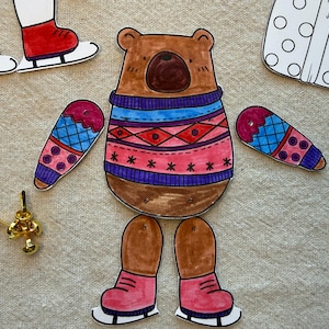Puede incluir: Un oso de papel con un su&eacute;ter con un patr&oacute;n azul y rojo, con mitones y patines de hielo a juego. El oso es un proyecto de manualidades, con piezas separadas para el cuerpo, los brazos y las piernas.