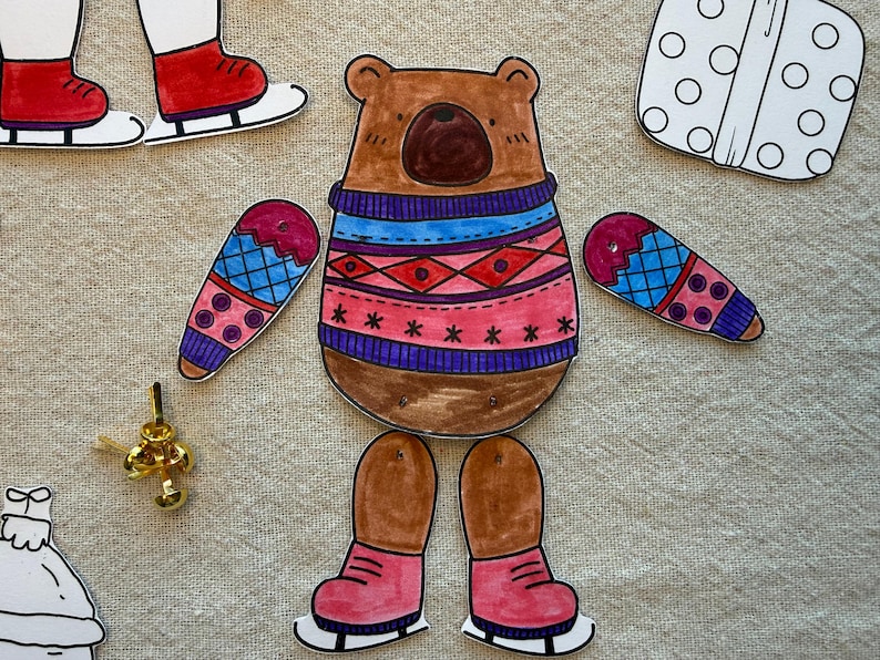 Puede incluir: Un oso de papel con un su&eacute;ter con un patr&oacute;n azul y rojo, con mitones y patines de hielo a juego. El oso es un proyecto de manualidades, con piezas separadas para el cuerpo, los brazos y las piernas.