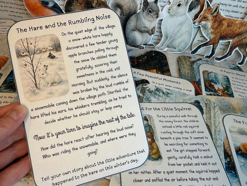 May include: A hand holds a card titled "The Hare and the Rumbling Noise" with a winter scene illustration. Other cards with animal illustrations and text are visible, suggesting a children's storybook theme.