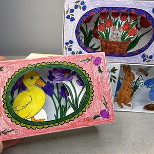 Puede incluir: Arte en papel dibujado a mano con un pato amarillo, un conejo marrón, un pájaro azul y un ratón en una cesta. El arte incluye flores y una abeja, todo dentro de escenas coloridas y enmarcadas. La obra de arte es de estilo diorama 3D.