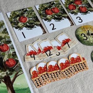 May include: Watercolor illustrations of apples and trees. The image includes cards with apple trees numbered 1, 2, and 3, along with apple basket cards numbered 3, 4, 5, 7, and 12. Green and yellow circular cards with dots and numbers are also present.