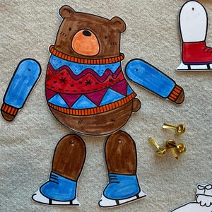 Puede incluir: Mu&ntilde;eca de papel de oso marr&oacute;n con un su&eacute;ter azul y rojo, brazos azules y piernas marrones con patines azules. Incluye chinchetas doradas para el montaje. Un divertido proyecto de manualidades para ni&ntilde;os.
