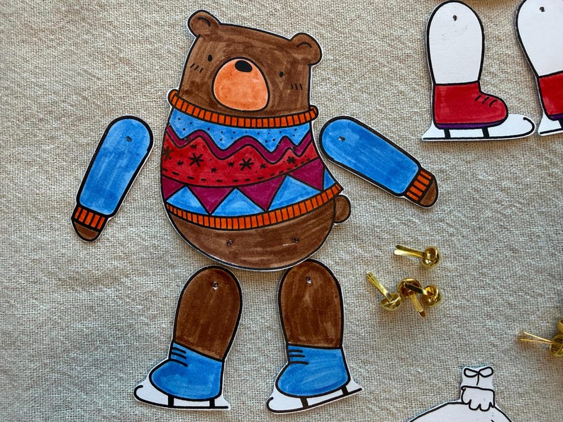 Puede incluir: Mu&ntilde;eca de papel de oso marr&oacute;n con un su&eacute;ter azul y rojo, brazos azules y piernas marrones con patines azules. Incluye chinchetas doradas para el montaje. Un divertido proyecto de manualidades para ni&ntilde;os.