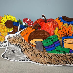 May include: A whimsical illustration of a hedgehog carrying autumn elements. The image features a sunflower, apple, pumpkin, mushroom, socks, and a mug of a warm beverage. The color palette includes orange, yellow, brown, and green.