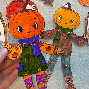 May include: Two paper dolls with orange pumpkin heads, each holding a lantern on a stick. One doll wears a green dress, purple scarf, and striped leggings. The other wears a brown sweater with maple leaf patterns, a green scarf, and boots.