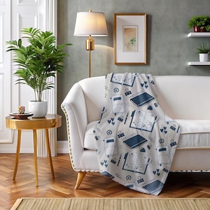 May include: A white throw blanket with a blue and white pattern of notebooks, pens, and paper clips. The blanket is draped over a white armchair in a living room setting.