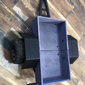 May include: A small, rectangular, gray utility trailer with a black hitch and black wheel fenders. The trailer features a hollow, open-top cargo bed with visible interior screws and two large, textured black rubber tires on the sides.