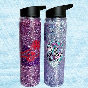 May include: Two purple glitter water bottles with different designs. One bottle has a red and white cheer design with the word "Queen" on it. The other bottle has a blue and pink dance design with a disco ball and smiley faces.