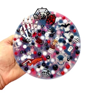 Puede incluir: Un disco circular de resina relleno de dijes con temática de Halloween, incluyendo una mano de esqueleto blanca, una telaraña negra, murciélagos, una lápida y una botella en miniatura etiquetada como Vampire Blood. Un dije de jugo dice Blood Juice.