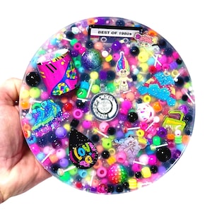 May include: A round, clear resin piece filled with colorful beads, charms, and trinkets. Includes a pink and blue geometric design, a Troll doll, and the text "BEST OF 1980s". The piece is held by a hand.