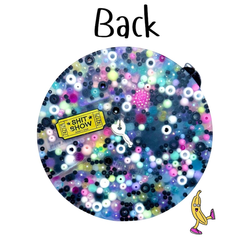 Puede incluir: Un llavero redondo y brillante con una mezcla de cuentas coloridas y un dije amarillo en forma de boleto que dice "SHIT SHOW". Tambi&eacute;n se ve un peque&ntilde;o dije blanco en forma de llave y un pl&aacute;tano de dibujos animados.
