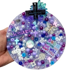 Puede incluir: Un adorno religioso redondo, de tono púrpura, con varios adornos. Presenta cruces, cuentas y la palabra "Jesus". El adorno también incluye el texto "God is within her, she will not fall."