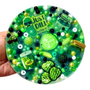 Puede incluir: Un posavasos redondo de resina verde con una variedad de cuentas verdes y gráficos con temática de pepinillos. El posavasos presenta imágenes de pepinillos, frascos de pepinillos y el texto "Just Dill With It."