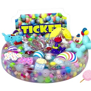 Puede incluir: Un cuenco de resina transparente lleno de cuentas de colores y figuras en miniatura con temática de circo. Incluye un elefante azul, un payaso, una foca y un animal de globo. Un letrero que dice "TICKET" está en el fondo, con un algodón de azúcar al lado.