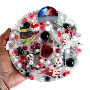 Pode incluir: Uma obra de arte redonda e transparente em resina com tema de cena de crime. Apresenta contas vermelhas, pretas e brancas, garrafas em miniatura, um martelo e um sinal que diz "DO NOT CROSS CRIMES SCENE".