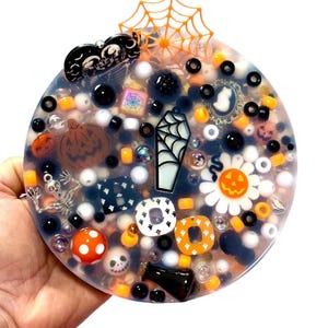 Puede incluir: Un agitador de resina circular lleno de abalorios y cuentas con temática de Halloween, incluyendo un ataúd, una calabaza, un esqueleto y un adorno de telaraña. La pieza presenta acentos negros, blancos y naranjas con varios motivos de fantasmas.