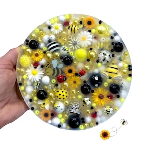 Puede incluir: Una pieza de arte de resina redonda con un tema de abejas. La pieza está llena de cuentas amarillas, blancas y negras, junto con adornos de abejas y flores. El diseño incluye girasoles, margaritas y mariquitas, creando una estética caprichosa inspirada en la naturaleza.
