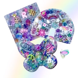 Puede incluir: Un accesorio de resina en forma de lazo colorido con una sirena, un arcoíris y otros elementos decorativos. El lazo está adornado con cuentas, flores y una pequeña ilustración de mariposa. La paleta de colores incluye tonos de morado, rosa y azul.