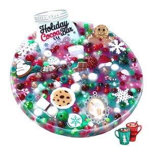 Puede incluir: Un slime circular transparente lleno de cuentas rojas, verdes y blancas, dijes de copos de nieve, un hombre de jengibre, una galleta y un letrero que dice Holiday Cocoa Bar. Contiene mini barras de chocolate, tazas y un marco ovalado.