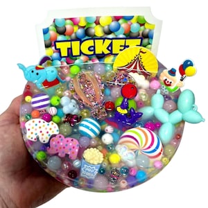 May include: A clear, circular resin art piece with a circus theme. It contains colourful beads, miniature circus-themed charms such as elephants, a hot air balloon, and a clown, and the word "TICKET".