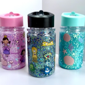 May include: Three clear plastic water bottles with colorful glitter and cartoon designs. The bottles have pink, black, and blue lids with a straw opening. The designs include a princess, sea creatures, and seashells.